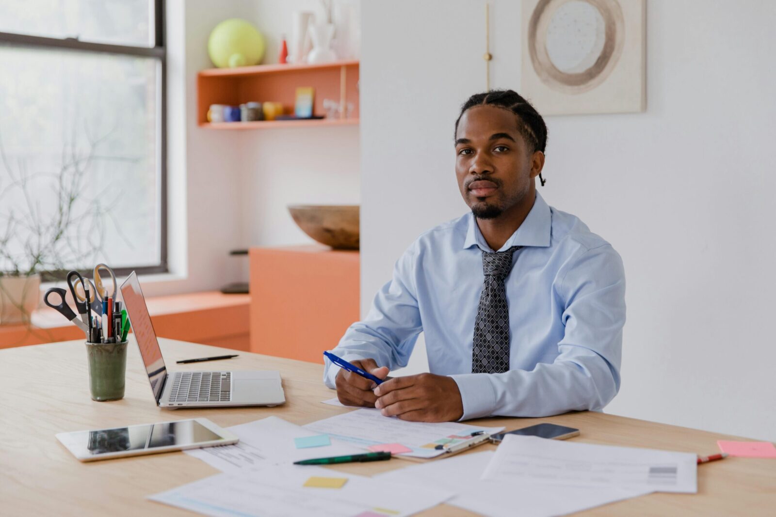A professional businessman with a beard and braided hair sits at his desk with a laptop and documents, working diligently.